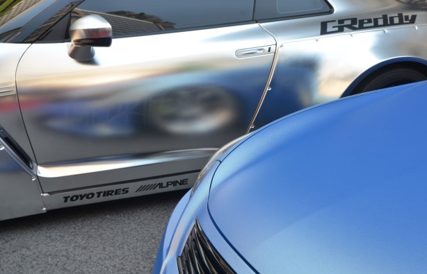 parts of two cars parked beside each other in a car show, one is blue and the other is silver. The front wheel of the blue car is reflected in the side panel of the silver car which has G Reddy painted by the rear window. 