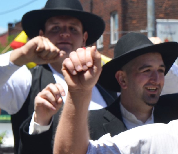 Close up shot of the hands of three men dancing in a parade. The faces of only 2 of them are visible. They are wearing white shirts, black vests, and black hats. Portugal Day parade