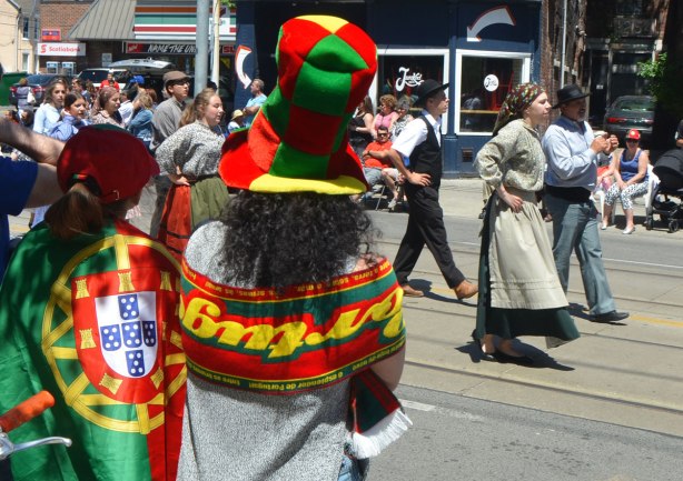 A young woman is wearing a large tall hat in squares of the colours of the Portuguese flag. Beside her is a person draped in the Portuguese flag. They are watching a parade