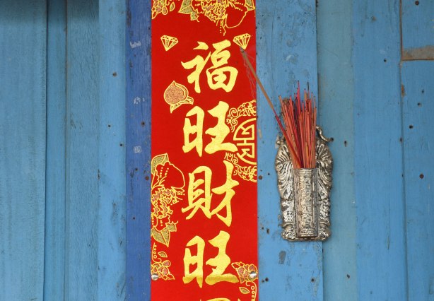 A blue wood door, a red banner with gold Chinese lettering, and a silver holder for incense sticks, many reddish incense sticks in it 