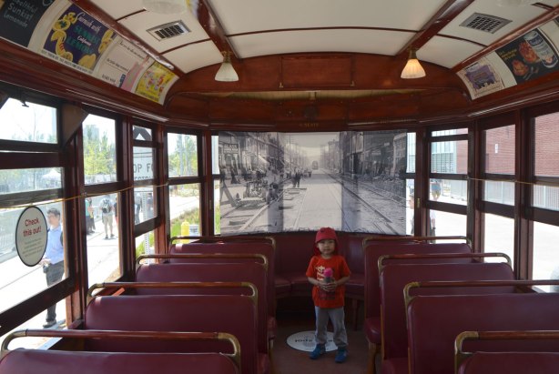 A young boy stands in the back of an old restored ttc streetcar. A black and white picture of an old street scene has been put across the back window to show you what the view out the window might have looked like at the time the streetcar was functional. Old ads on the upper part of the interior, wood trim