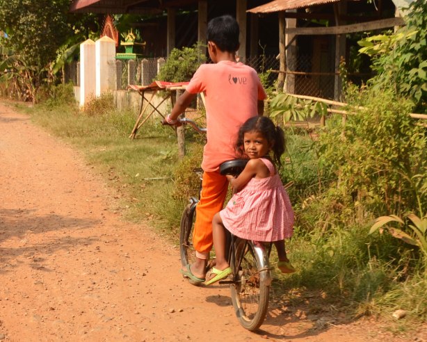 a young girl sits on the back fender of a bike ridden by a boy. She is wearing a pink dress and she is looking back at the camera. The boy is dressed in orange and only the back of him can be seen 