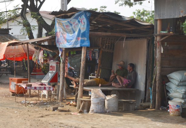 Men rest in the shade of a makeshift shelter beside the road. 