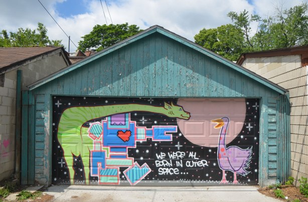 mural on a garage door in an alley. a green giraffe, a blue lovebot, and a purple goose, with the words "We were all born in outer space"