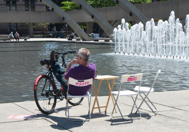 A man sits on a folding chair beside the fountain at Nathan Phillips Square. He has his feet up on the bars of his bike that is balanced there.