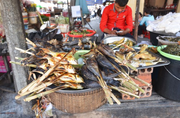 A young man in a red shirt looks at his phone while he sits behind some cooked fish on sticks that he is selling at a market