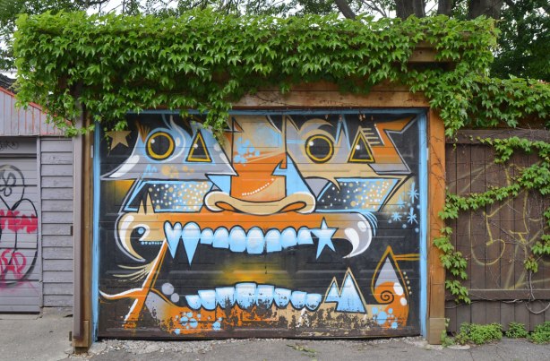 garage door painted with a monster face, big mouth and teeth, orange nose, in tones of blue, grey and orange, ivy is growing over the top of the garage and it looks like green hair. 