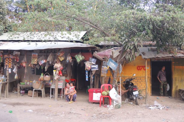 a woman squats beside her young daughter in front of a store that is selling many things that are hanging from the overhang on the exterior of the store