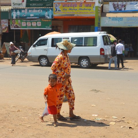 a mother and son, both dressed in orange clothes, walk along the side of a road. A white van is in the background. 