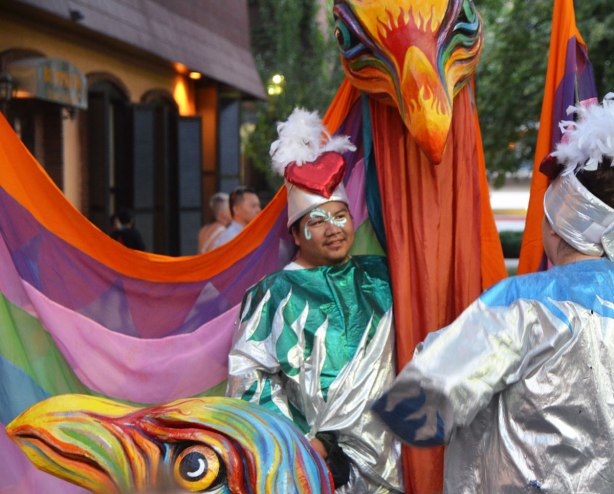 a man holding a large pole with a bird head on the top of it, with rainbow coloured fabric, meant to be the bird's wings. 