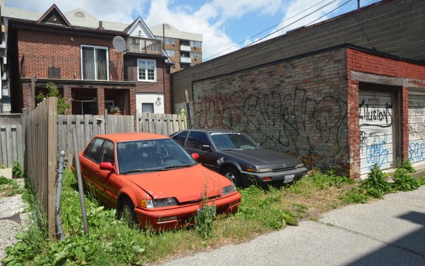 two old cars, one red and one dark grey, are parked in an overgrown backyard on an alley.