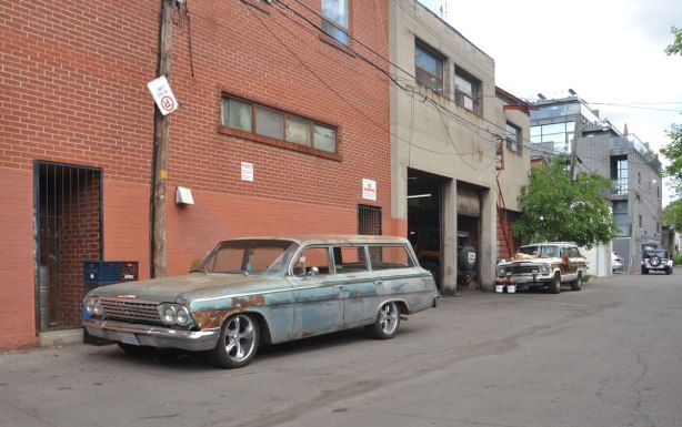 wide lane behind Dupont Street in Toronto, an old car is parked there, back of an auto repair shop, sturdy looking two storey brick buildings. 