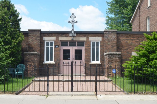 low, one storey brick building with a pink double door, metal fence and gate in front, ornate cross above the door.