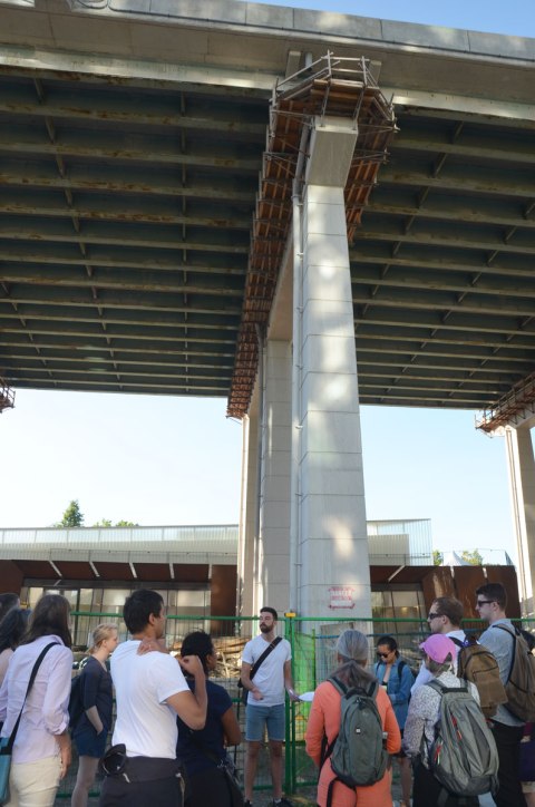 a man is standing in front of a group of people on a walking tour, he is standing under the Gardiner Expressway where it is 5 storeys above ground level. 