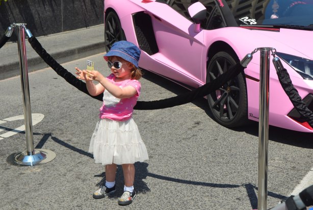 A little girl in white skirt, pink T-shirt, blue hat and big sunglasses stands in front of a fancy pink car at an outdoor car show