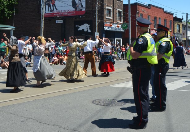 Two Toronto policemen in yellow safety vests watch a parade, women dancing past them.