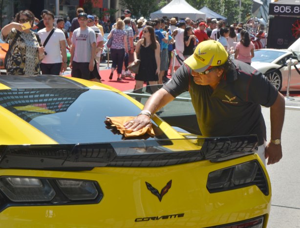 a man in a black short sleeve shirt and a yellow baseball cap is cleaning and polishing the rear window of a yellow Corvette at a car show, lots of onlookers in the background. 