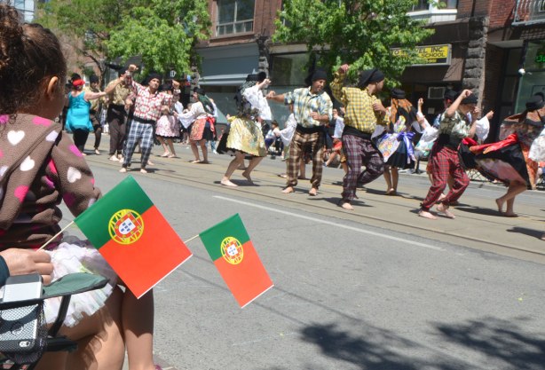 Spectators in a parade hold small Portuguese flags as they watch traditional dancers perform.