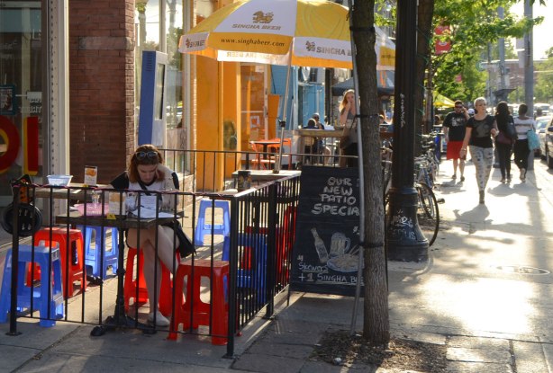 a woman sits on an outdoor patio, under a large yellow and white umbrella with a Singha Beer ad on it, sitting on a bright red plastic stool. Other plastic stools are around her, both red and blue
