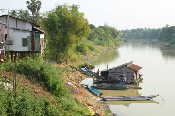 river, on the riverbank is a house on wood stilts, at river level is another house that is floating on the water. Two flat boats are docked by it. 