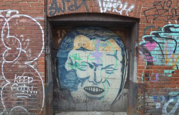 an old doorway in a red brick building in a lane. There is an old graffiti face of Rob Ford painted on the door. R I P has been written on his forehead. 