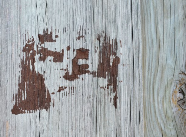 a brown stencil of a man's face on a wood fence but the paint was a bit heavily applied and there are some paint blobs 