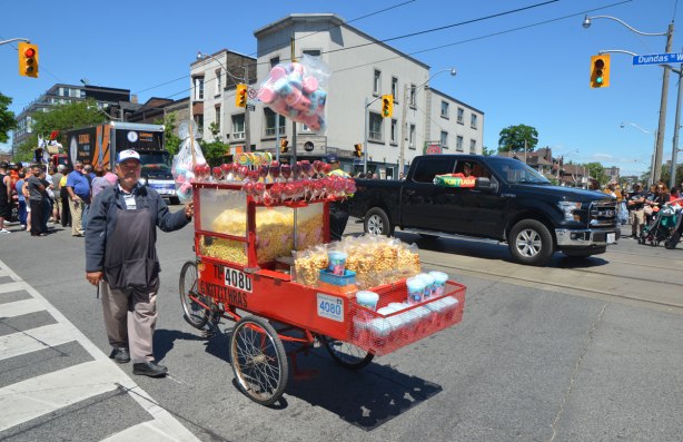 A man pushes a cart from which he is selling popcorn, candy apples and cotton candy to people watching a parade. A black truck is behind him with a boy in the passenger side who is hold a banner out the window that says Portugal on it.