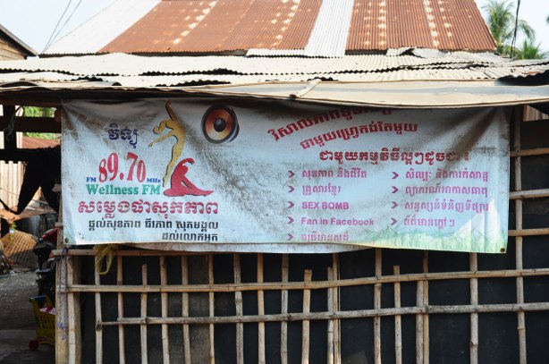 a banner outside a rough wood building, an ad for a Cambodian radio station 89.70, Wellness FM, 