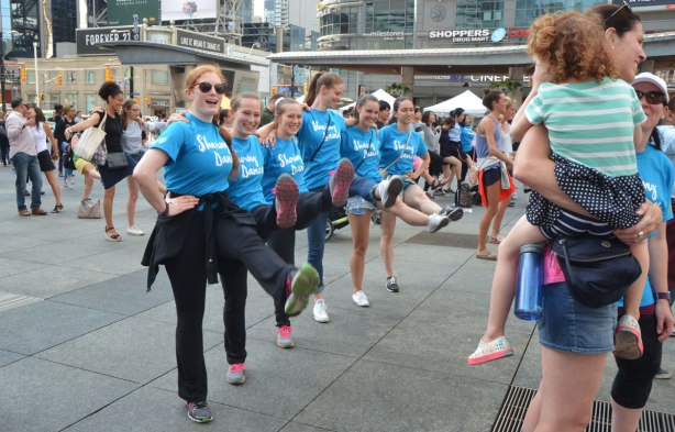 people dancing at Yonge Dundas Square as a group, part of an event called Sharing Dance - a kick line of 5 or 6 young women