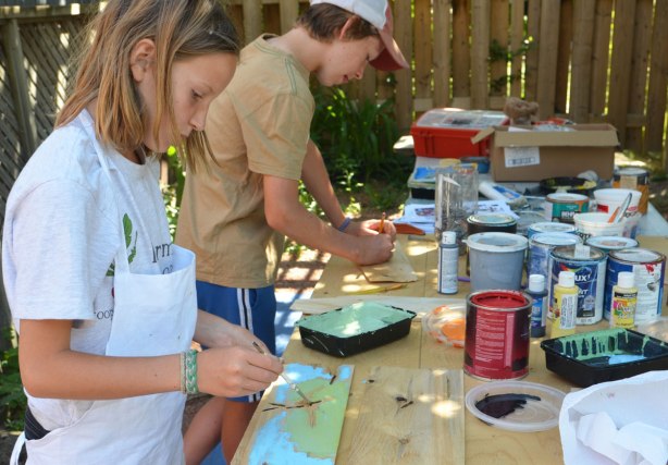 kids painting designs on wooden shingles, outdoors.