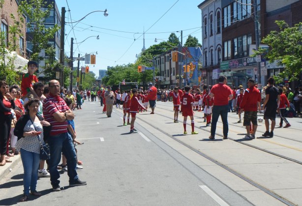 people watching kids kicking around a soccer ball as part of a parade. The kids are all in red uniforms.