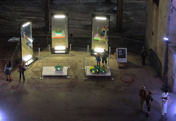 looking down onto the ground floor of the hearn generating station at luminato festival, three large vertical mirrors and some round mirrors on two tables. A few people looking at the mirrors, some other people standing around. 