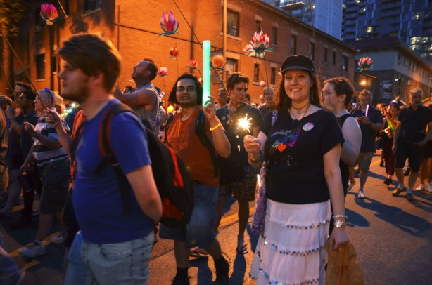 people walking in a night time parade for nuit rose, down Church St., one man is holding up a light stick, a woman is holding a sparkler, other people have lanterns and glow sticks. 