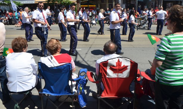 Spectators at a parade, sitting on chairs, one of which has a Canadian flag on the back. Passing by is a marching band in dark pants and white shirts. Portugal Day parade on Dundas West.