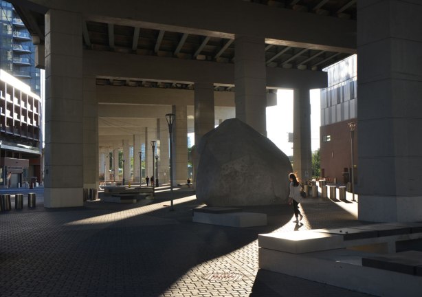 evening, low angle sunlight shines under the Gardiner Expressway where it has been developed with paving stones and some large rocks. 