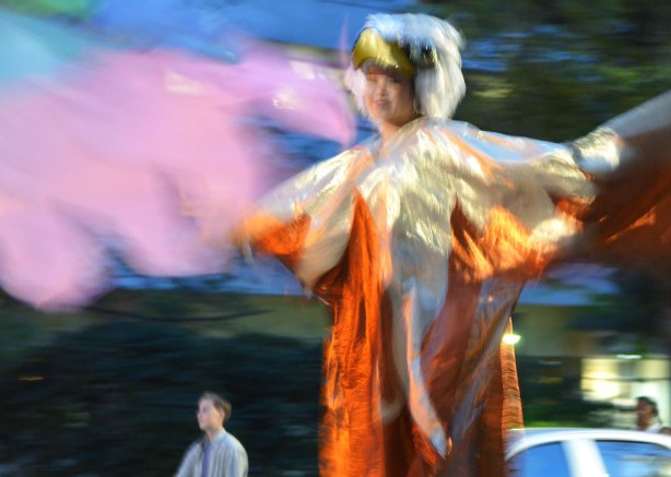 A woman in a flowing costume with eagle head, up on stilts, in a night time parade for nuit rose