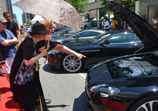 a man with grey hair and black glasses is pointing to the front of a black sports car with its hood up. He is pointing out something to the woman beside him who is dressed in black with black hat and holding an umbrella over her head. They are behind a barrier at an outdoor exotic car show