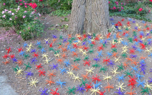 under a tree in a park, with roses in the background, many coloured 3D stick shapes that have been covered with yarn, lie on the ground. 