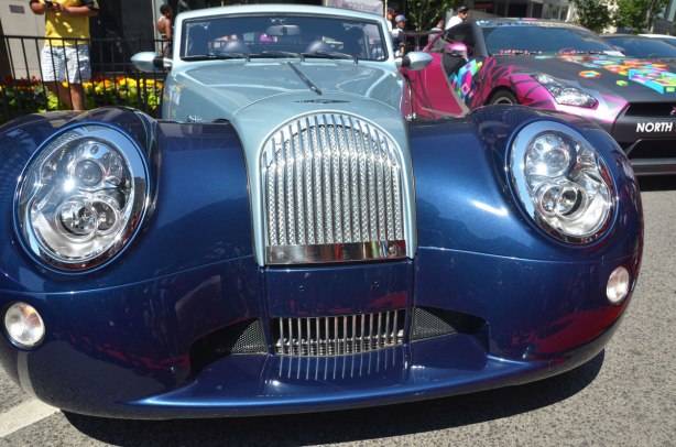 front end of a two toned blue Morgan car in a car show outside on Bloor Stree, with a rally car in different colours beside it. 