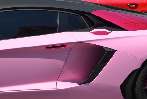 the side of a pink lambourghini, bousoughini, with a reddish car behind it. 