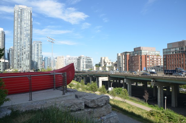The end of a large red canoe, an artwork by Douglas Coupland called 'Tom Thomson's Canoe' sits in a park, high above the surrounding scenery. Looking southwest towards the elevated Gardiner Expressway and the condo developments south of it. 