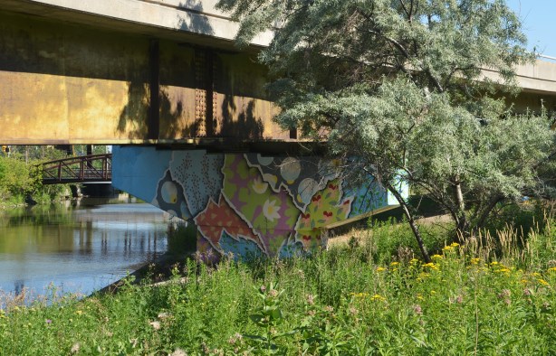 a bent supporting an offramp has been covered in a bright mural, grass and weeds grow in front, the river is behind, a small tree also in the picture 
