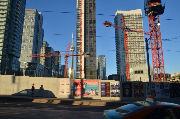 late afternoon, long shadows, yellowish tint to the photo, looking east from Bathurst, south of Front, north of the Gardiner, wood construction hoardings with posters on it, many orange cranes, some condos already built, a woman jogging past, cars on the street. 