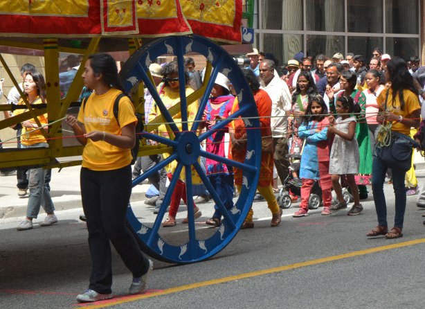 a large blue wheel that is holding up a chariot float in the Festival of India parade, people walking beside and behind it as they walk down Yonge Street