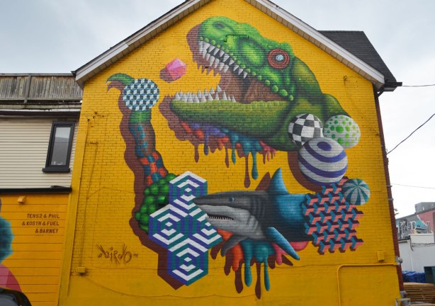 large mural by birdo, street art, on a two storey bright yellow building, a shark, and an alligator (or crocdile) head 