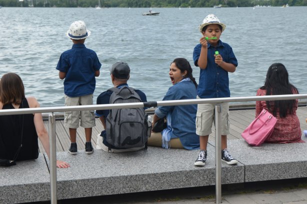A family sits by the lake, two young sons in matching fedoras. Mother is yawning, father and one son have backs to camera, other son is blowing bubbles towards the camera