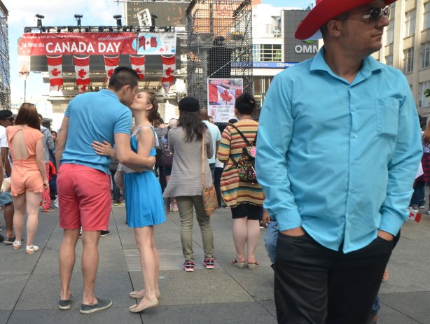 A man in a red cowboy hat in the foreground on the right, a couple kissing on the left. People watching a show on a stage in the background with Canadian flags and a banner that says Happy Canada Day