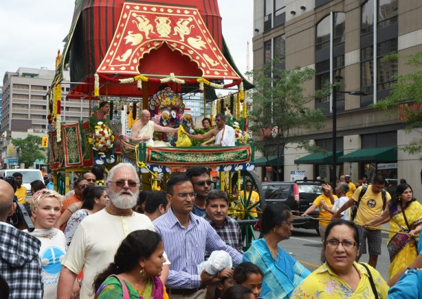 some older people dressed in white riding high in the chariot float in the Festival of India parade, others walking in front and pulling ropes to make the chariot move.