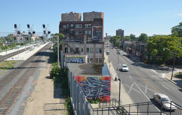 view from a bridge, a street, and a railway and some buildings in between. There is a mural at the bottom of the steps. 