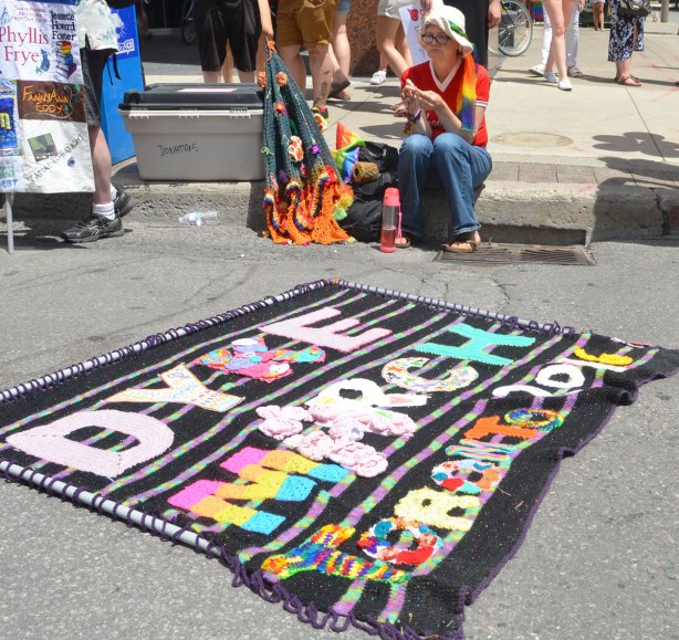 crocheted banner for dyke march is lying on the pavement. A woman is sitting on the kerb and she is crocheting